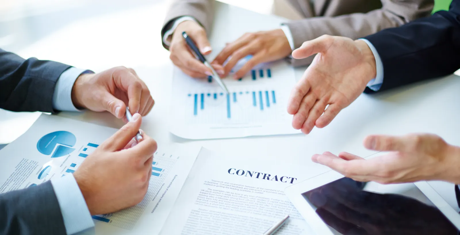Close-up of business professionals discussing charts and graphs during a meeting, with documents, a contract, and a digital tablet on the table.