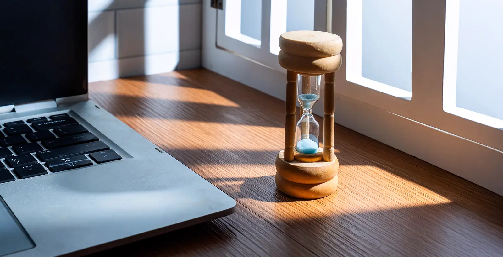 An hourglass beside a laptop on a sunlit desk, symbolizing SOC 2 report validity and renewal timelines.